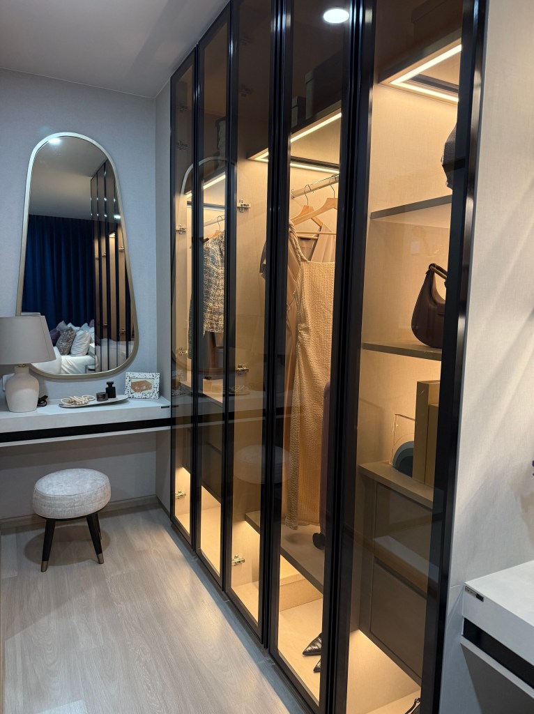 Interior view of a modern wardrobe featuring glass doors, illuminated shelves displaying clothing and accessories, and a small dressing area with a stool and mirror.