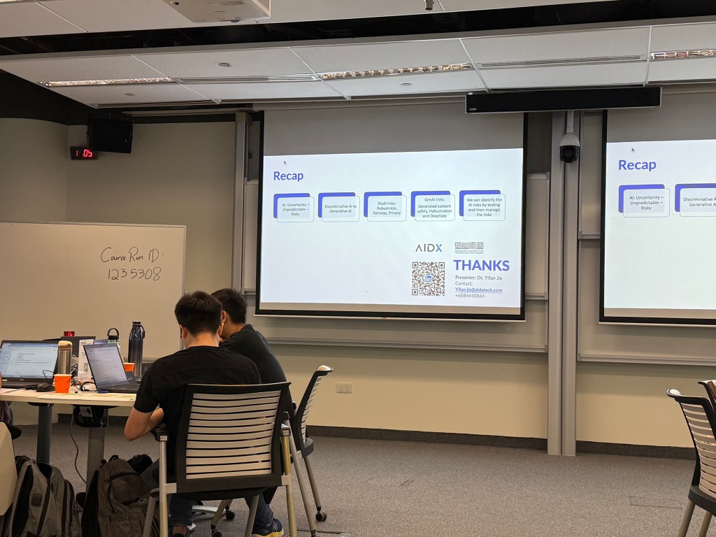 A classroom setting with students seated at tables, engaging with laptops. A projector screen displays notes on AI risks and the importance of careful management. The environment suggests a learning atmosphere focused on artificial intelligence education.
