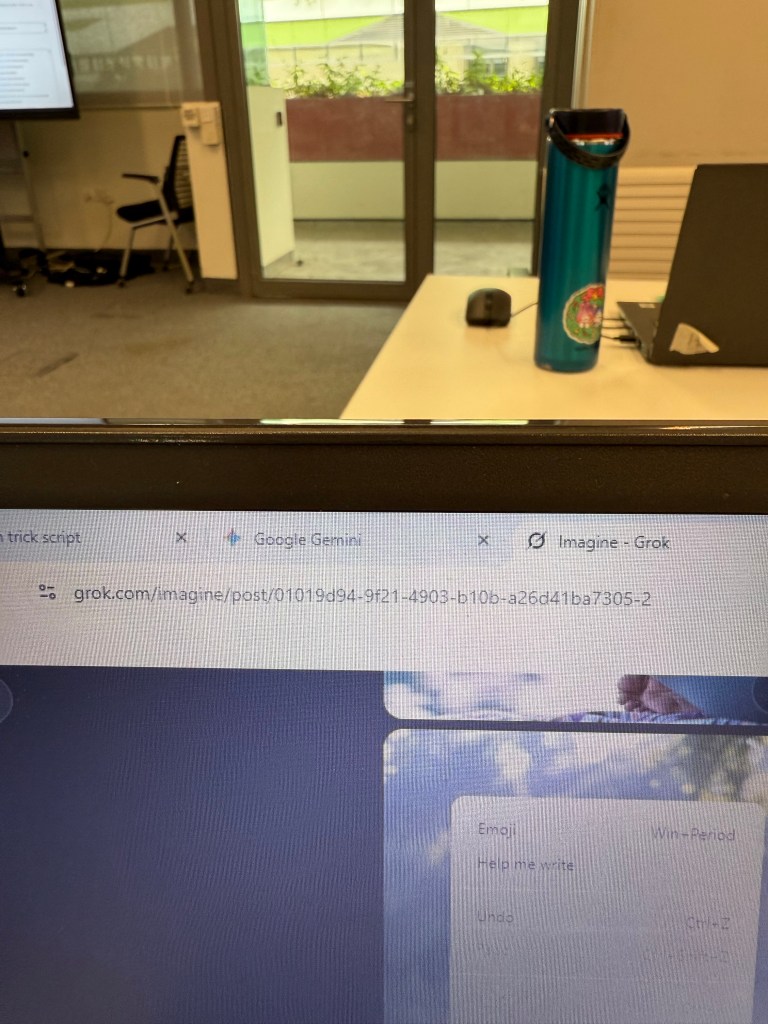 An interior view of a workspace featuring a laptop in the foreground, a blue water bottle, and glass doors leading to an outdoor area. A screen displays content related to a digital program.