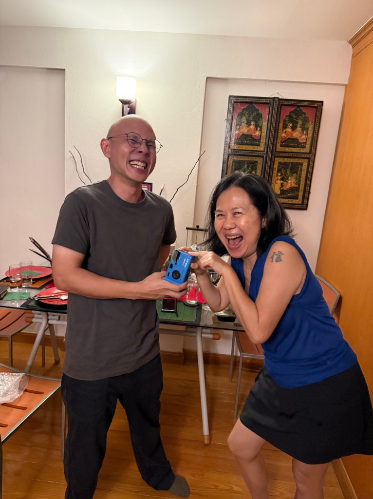 Two smiling adults, one holding a gift camera while the other joyfully reacts, in a warmly lit indoor setting with wooden flooring and artwork on the walls.