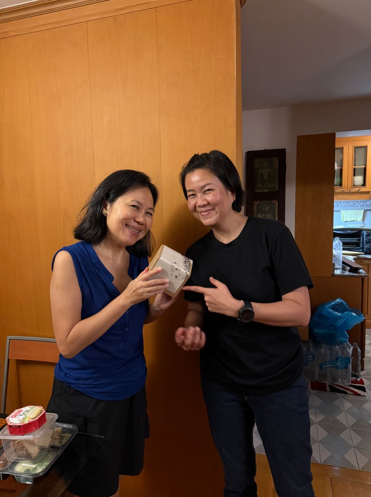 Two women smile together while holding a small gift box in a warm, homey setting.
