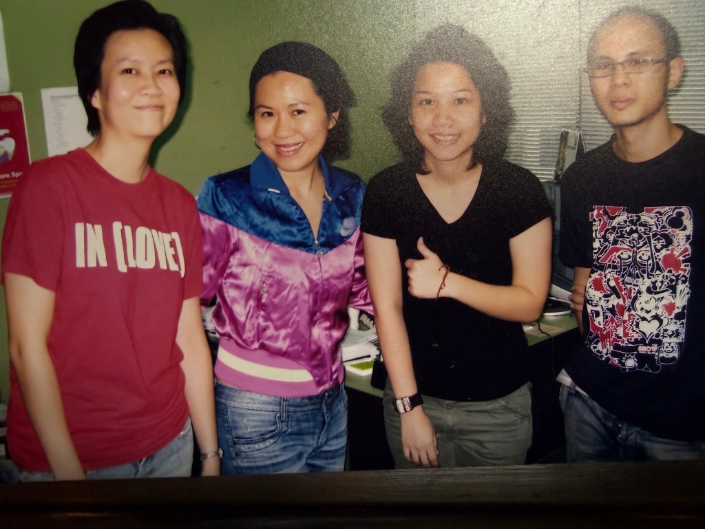 A group photo of four individuals smiling together in a casual setting, with desks and office supplies visible in the background.