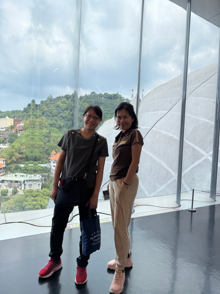 Two women standing together in front of a large window with a scenic view of green hills and cloudy sky.