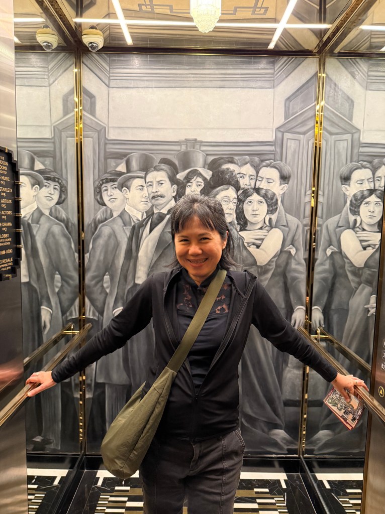 A woman smiling in an elevator, with a vintage mural of people dressed in early 20th-century clothing in the background.