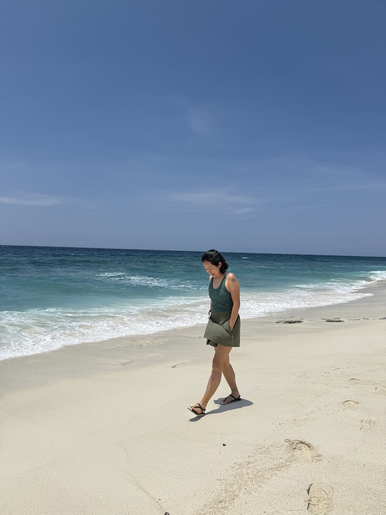 A candid, full-body shot of a woman walking peacefully along a sun-drenched white sand beach with turquoise waves, symbolizing the clarity and groundedness found when choosing the "messy truth" of confrontation over the heavy burden of avoidance.