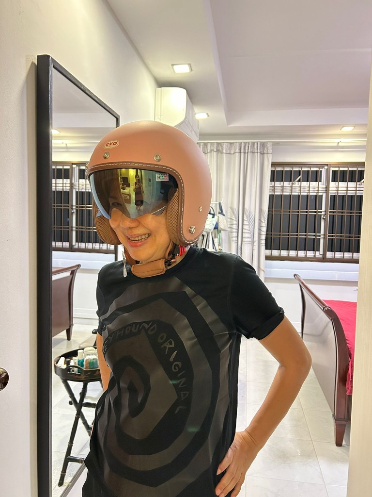 A smiling person wearing a pink helmet with a visor and a black t-shirt featuring a spiral design, standing in a bright indoor space.