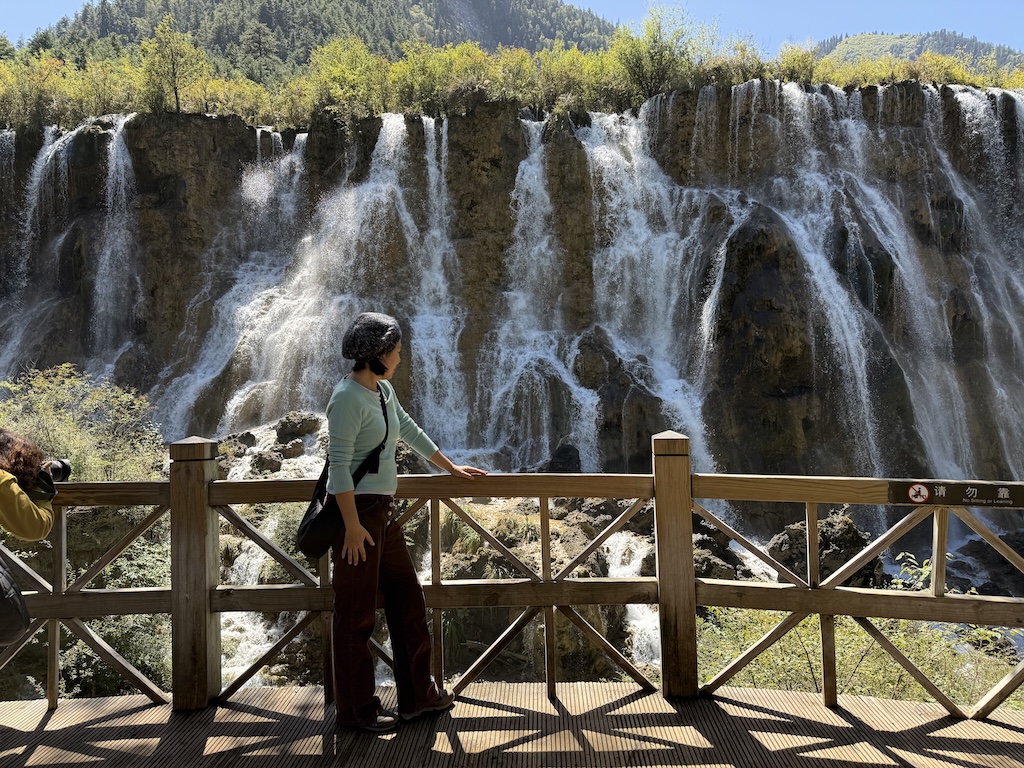 Blogger standing on a wooden bridge overlooking the turquoise Five Flower Lake in Jiuzhaigou National Park, China.