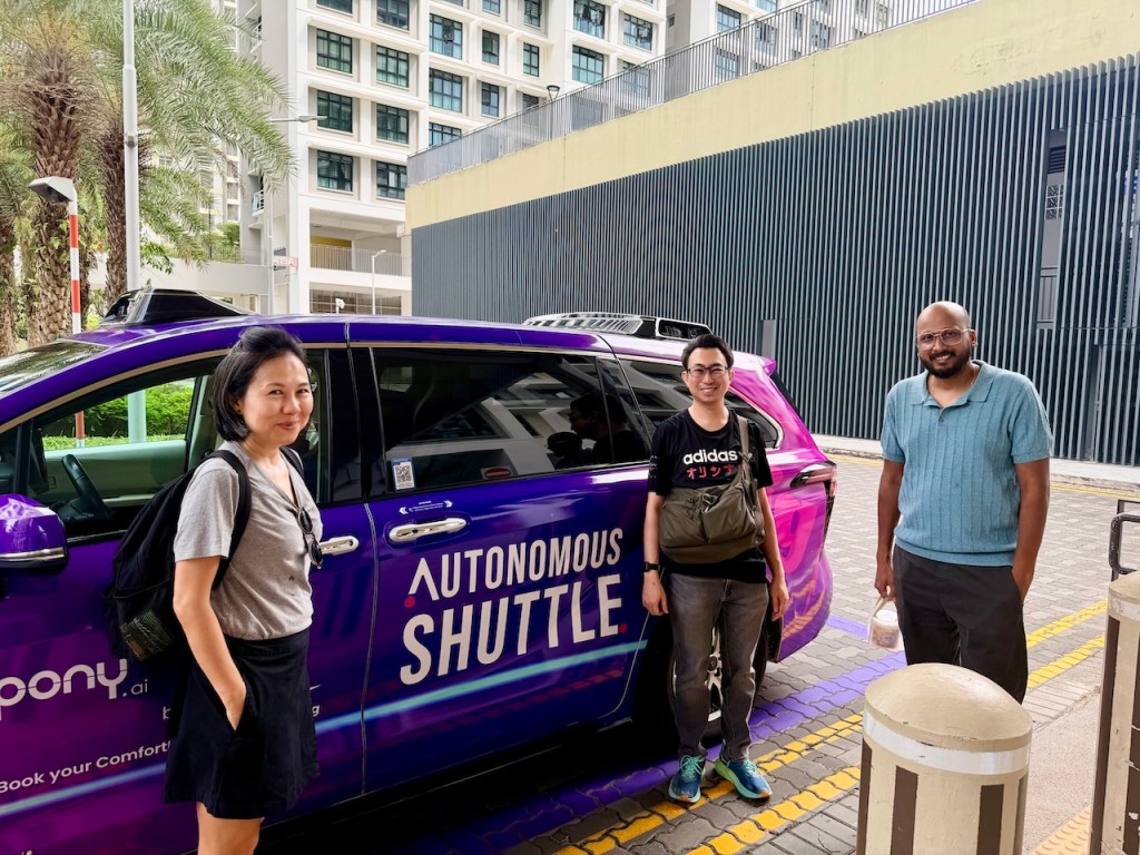 Jane Bay and two other "Future Riders" smiling and posing next to the side of a purple autonomous shuttle vehicle.