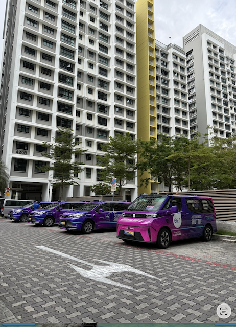 A fleet of purple and blue autonomous shuttle vans parked in a row in front of modern HDB apartment buildings in Punggol.