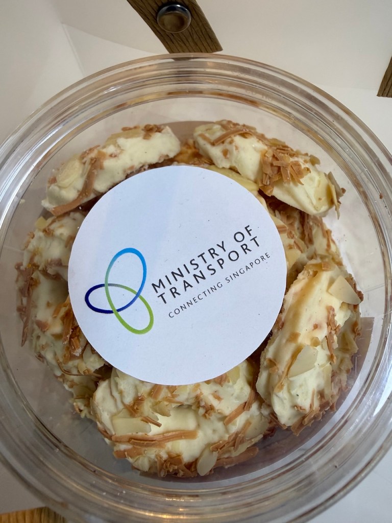 A clear container of cheese cookies with a white sticker featuring the Ministry of Transport Singapore logo.