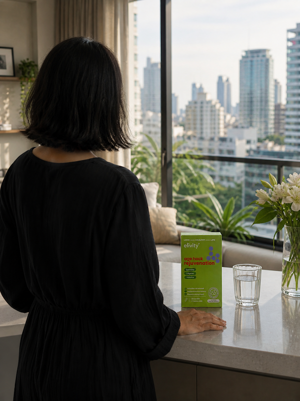 A woman in a black dress viewed from behind, looking out a window at a city skyline with an elivity Age Hack Rejuvenation box and a glass of water on the counter in front of her.