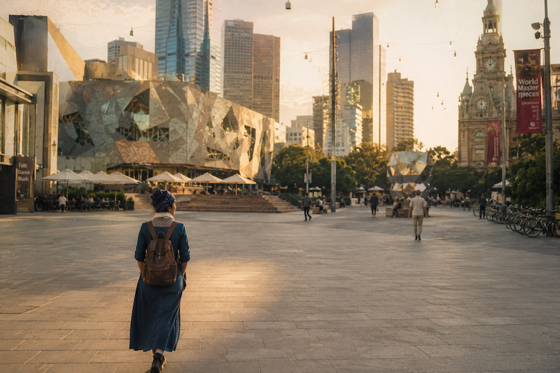 A vintage photograph from behind of a solo female traveller in a denim dress and backpack, walking through Melbourne's Federation Square at golden hour. The image has an analog film look and a digital timestamp that reads 'SEP 86'.