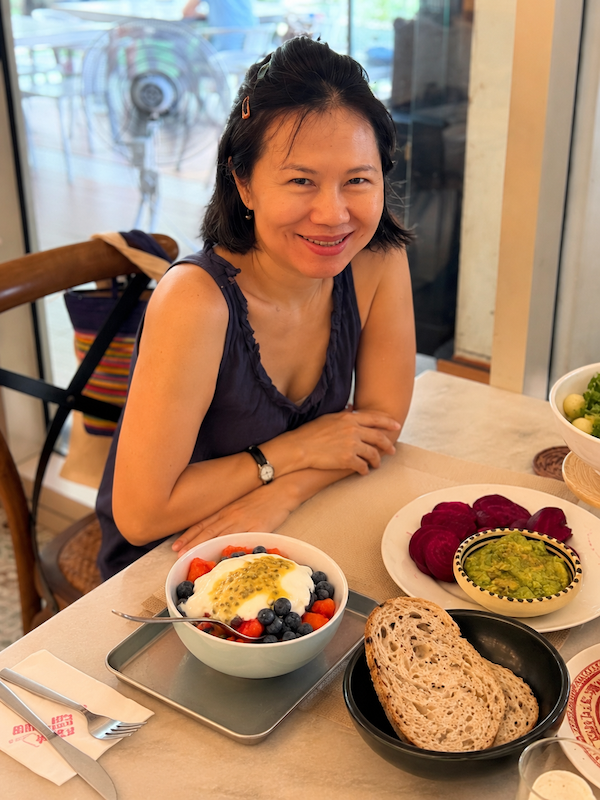 Active Aging blogger Jane smiling at her dining table with a gut-healthy brunch bowl of Greek yogurt, berries, and seasonal fruit.