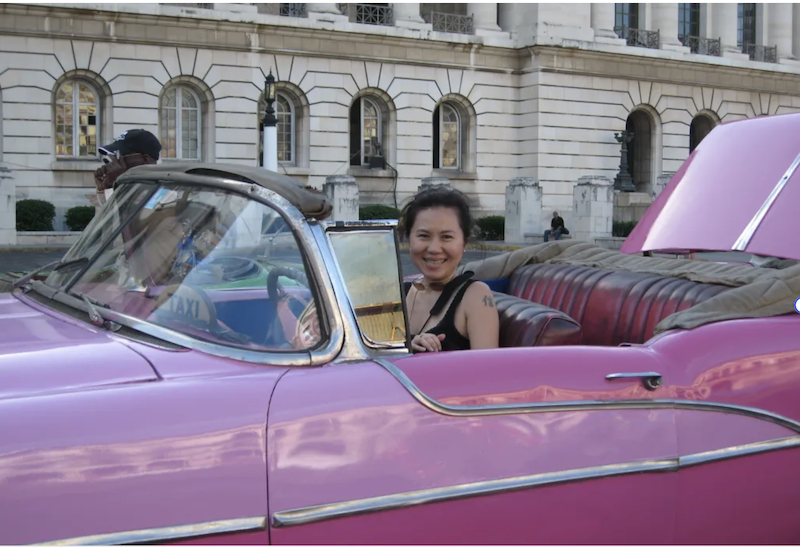 A smiling woman enjoying a ride in a classic pink convertible car in Havana, Cuba, with historic white stone buildings in the background.
