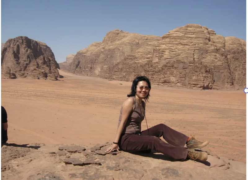 A solo female traveler sitting on a rock overlooking the desert landscape of Wadi Rum in Jordan. She is wearing hiking boots and smiling, with massive red sandstone rock formations in the background under a bright blue sky.