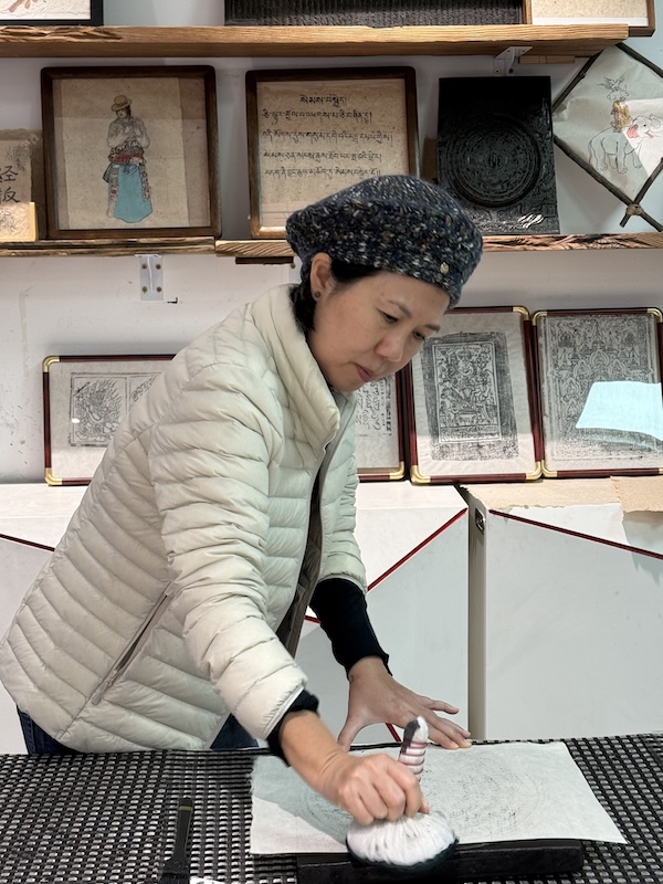  A woman wearing an Anta beige down feather jacket and a knit beret, carefully making a traditional Tibetan paper rubbing at a cultural art and craft workshop near Hotel Indigo in Jiuzhaigou.  