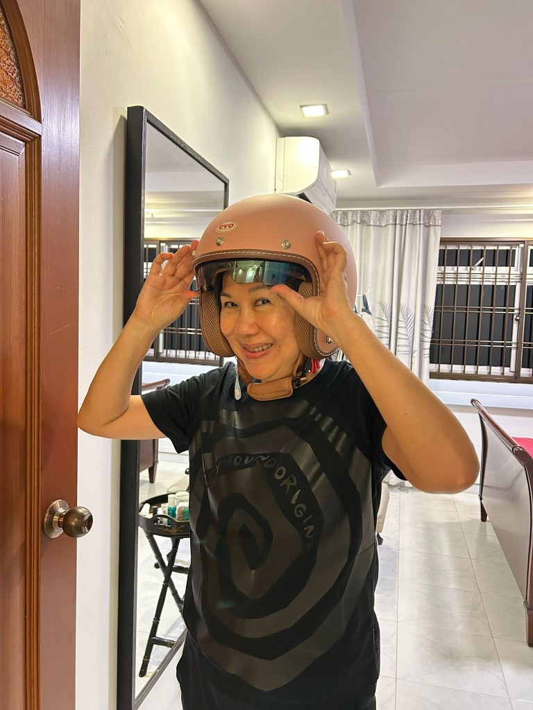 A woman smiling indoors while adjusting the visor of a stylish pink and tan Evo open-face motorcycle helmet.