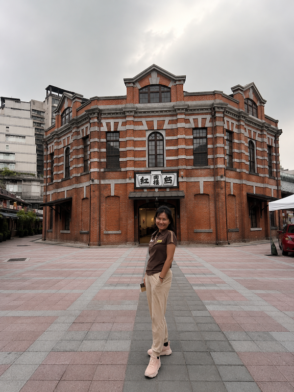 A woman standing in a stone plaza outside a historic red brick building, smiling while wearing comfortable Anta beige pants and pink Duozuolu walking shoes.