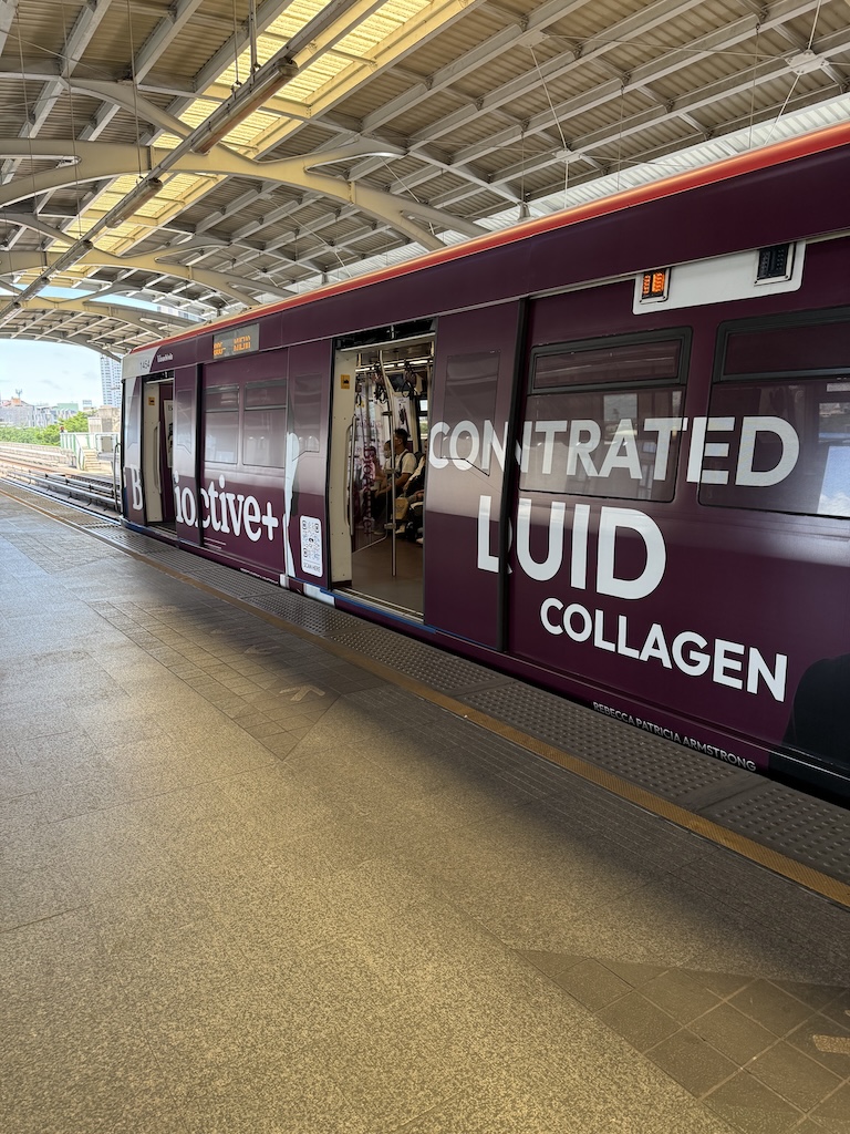 A deep maroon Bangkok BTS Skytrain stopped at an elevated open-air platform, featuring a prominent side advertisement wrap for bioactive liquid collagen.