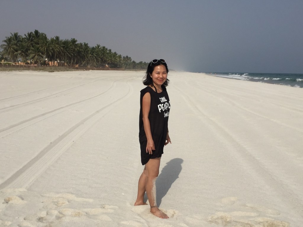 Jane standing on the white sand beach in Salalah, Oman, reflecting on the value of travel experiences over financial wealth.