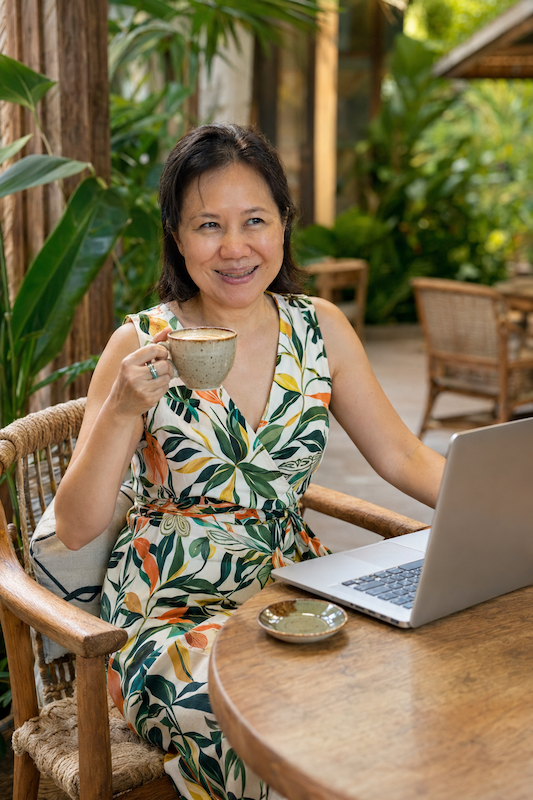 A youthful Asian woman in her 50s sitting in a cafe, looking thoughtfully away from her laptop with a cup of coffee.