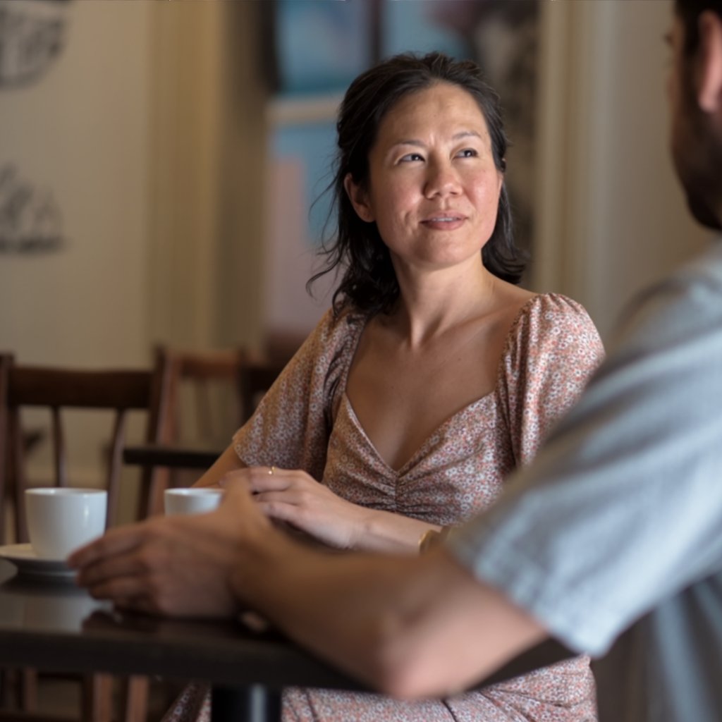 A mature Asian woman in her 50s speaking calmly and respectfully face-to-face across a café table.
